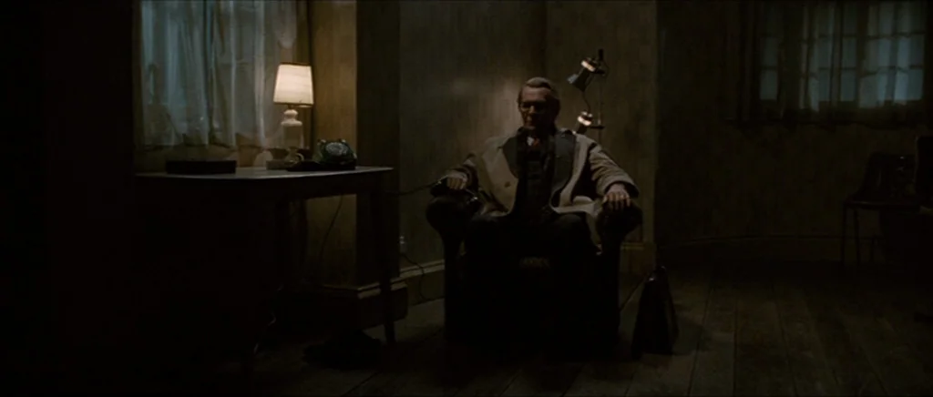 Tinker Tailor Soldier Spy (2011) movie still: wide — A dimly lit room contains a wooden table, armchair, and a telephone. An elderly man sits in the…