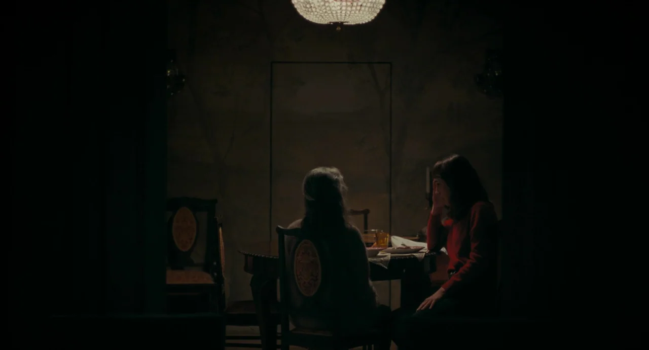 La Abuela (2021) movie still: wide — A dimly lit room with wallpaper featuring faint tree silhouettes has a dark wood table. Two women are seated…