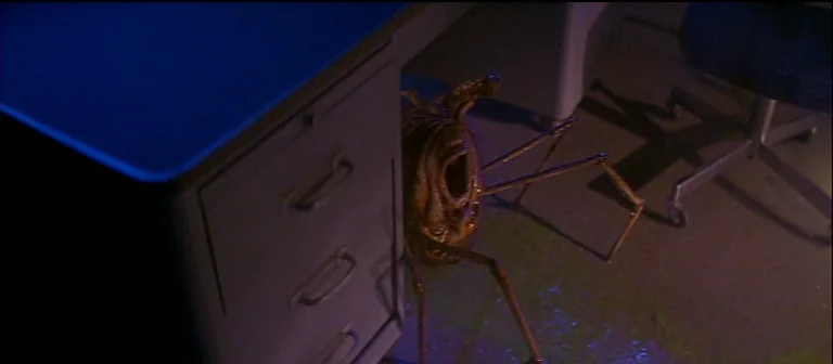 The Thing (1982) movie still: medium — A dark, utilitarian work area contains a metal desk with blue-topped surface. A spindly, organic-textured…