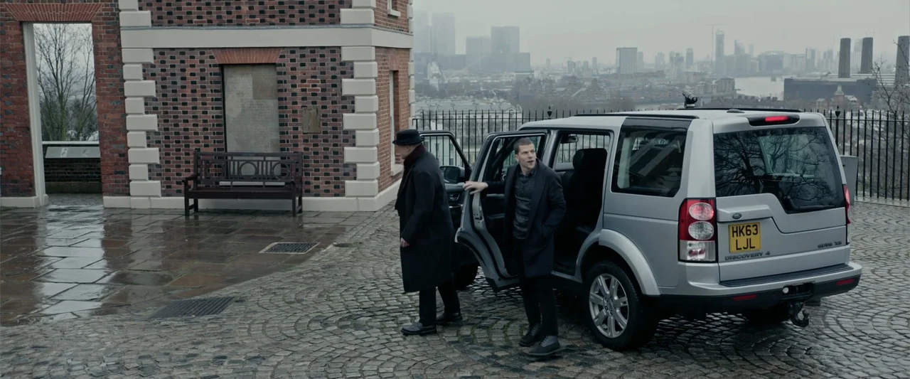 Now You See Me 2 (2016) movie still: wide — A wet cobblestone courtyard with a brick building and a silver SUV parked outside. A younger man steps…