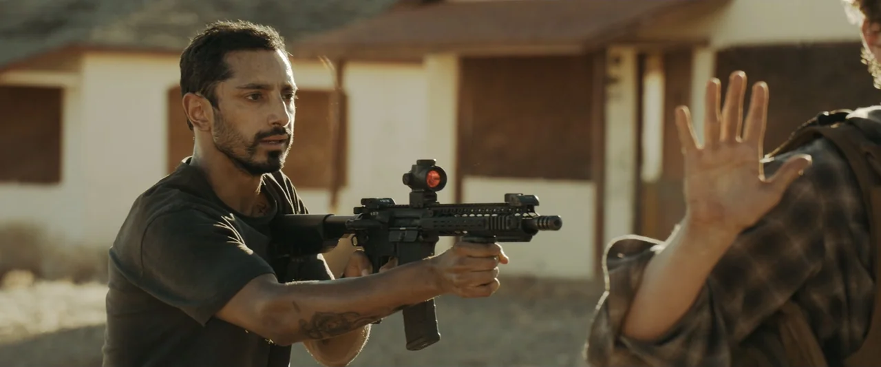 Encounter (2018) movie still: medium — A dusty outdoor environment features a blurred low building in the background. A man holds a tactical rifle…