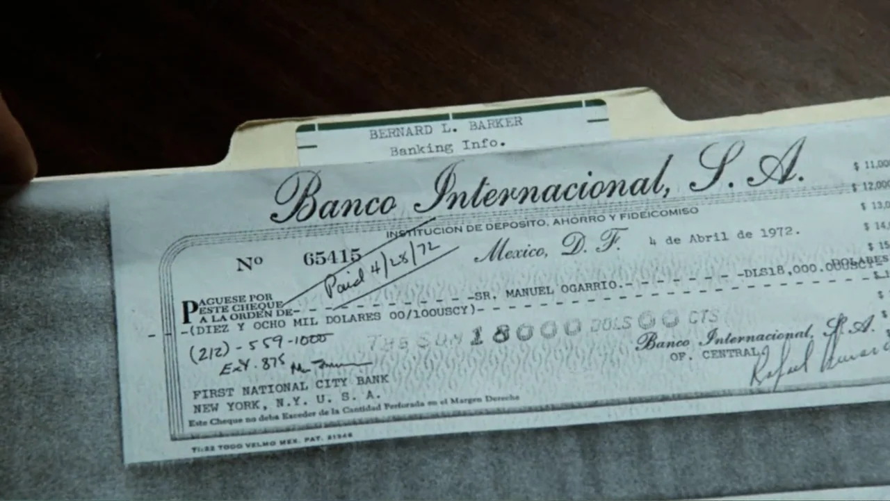 All the President's Men (1976) movie still: closeup — A manila folder sits on a dark wooden surface holding a bank document. The check details the…