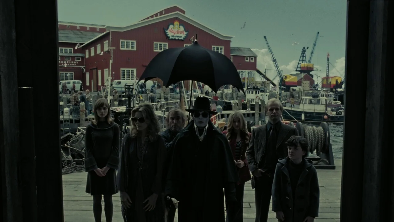 Dark Shadows (2012) movie still: wide — A group of six people stands on a weathered wooden pier by the water. A large red industrial building with a…