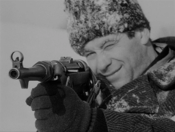 The Ascent (1977) movie still: closeup — A man stands in a snowy environment with frost visible on his hat. He aims a submachine gun forward with…