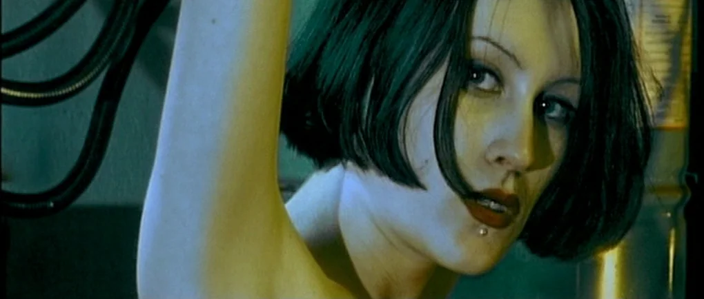 Mortal Transfer (2001) movie still: closeup — A close-up of a woman stands in front of industrial equipment with metallic textures. She looks at the…