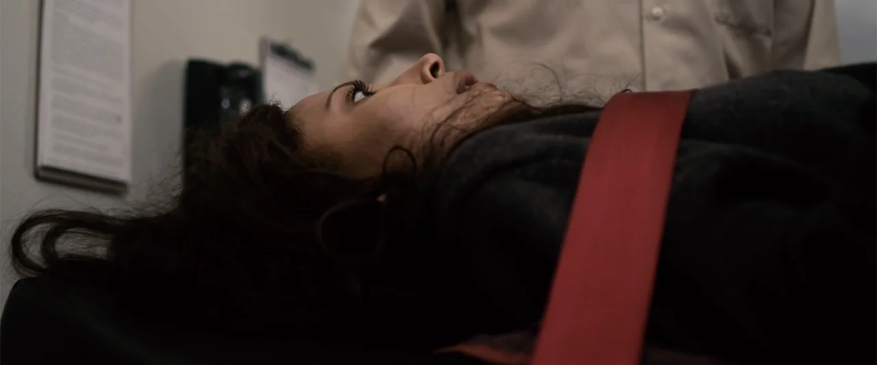 Mermaid Down (2019) movie still: closeup — A stark medical examination room features plain walls and clinical equipment. A young woman lies supine…