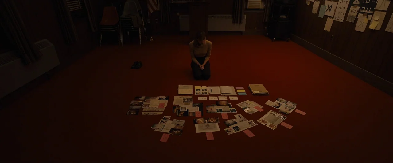 Longlegs (2024) movie still: wide — A dimly lit room features a large red carpet covered in scattered investigation documents. A young woman kneels…
