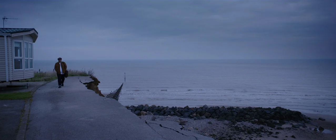 Ghost Stories (2017) movie still: wide — A man walks along a damaged asphalt path leading to an eroded coastal cliff edge. Beside him stands a…