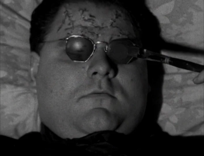 Tales from the Gimli Hospital (1988) movie still: A man lies prone with his eyes concealed behind dark spectacles as a mysterious, unidentified…