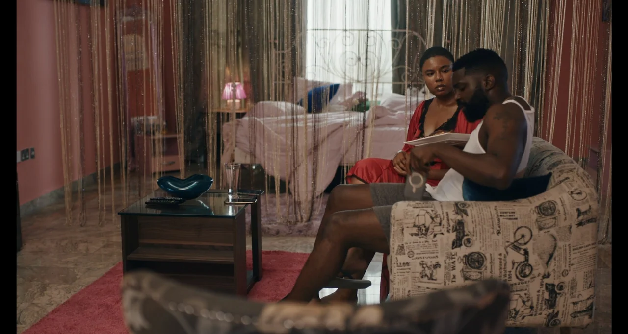 Love In A Pandemic (2023) movie still: medium — A furnished bedroom contains a central armchair and a bed draped in beaded curtains. A man sits…