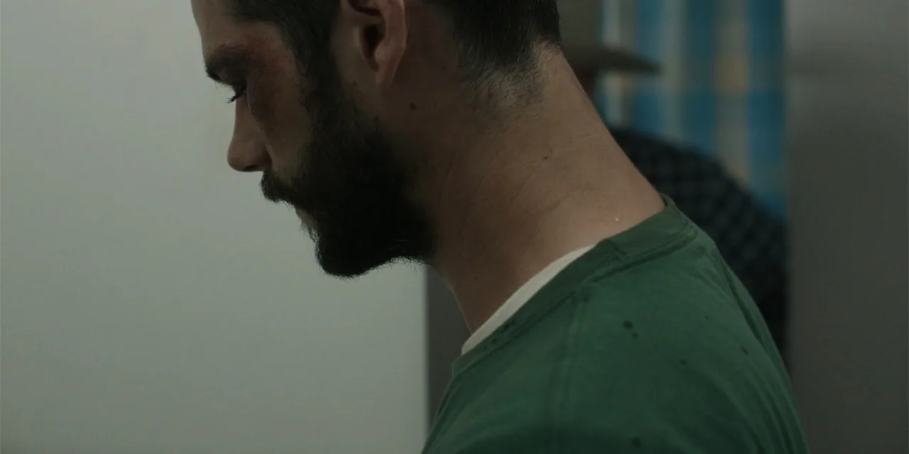 Caddo Lake (2024) movie still: closeup — An interior space with a plain wall and a hint of a curtain. A man with visible facial bruising and a beard…