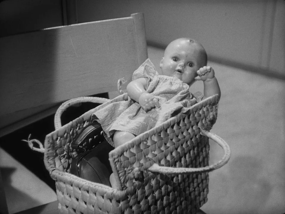 Brink of Life (1958) movie still: closeup — A vintage doll rests in a square woven basket placed on a chair. The basket also contains a small dark…