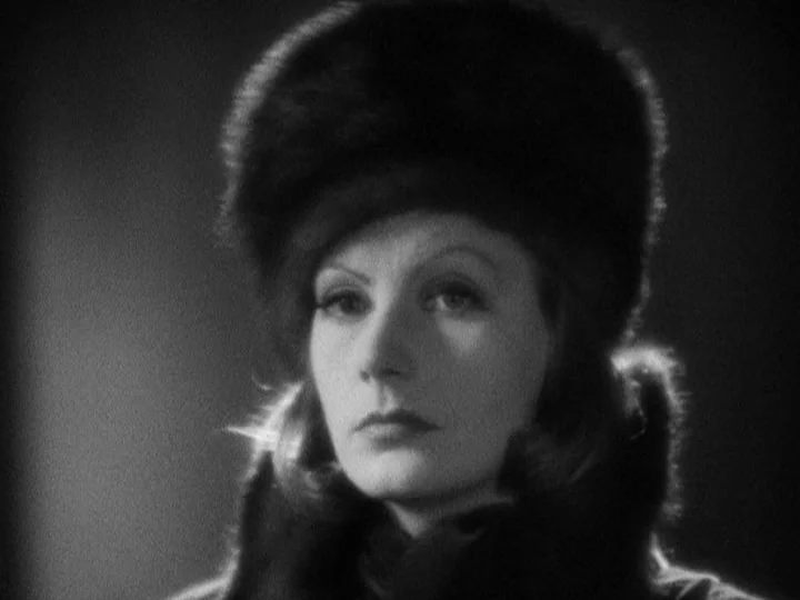 Queen Christina (1933) movie still: closeup — A close-up shot of a woman against a dark, featureless backdrop. She wears a thick fur hat and…