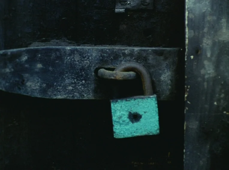 Daisies (1966) movie still: closeup — A heavy metal hasp is secured to a dark, weathered door with a rusted padlock. The turquoise-colored padlock…