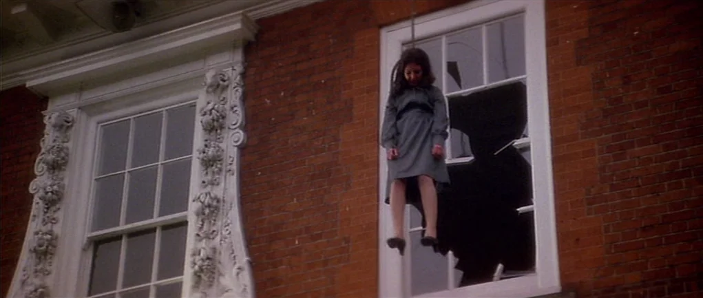 The Omen (1976) movie still: fullbody — A red brick wall with ornate white trim contains a shattered window frame. A girl in a grey dress hangs…