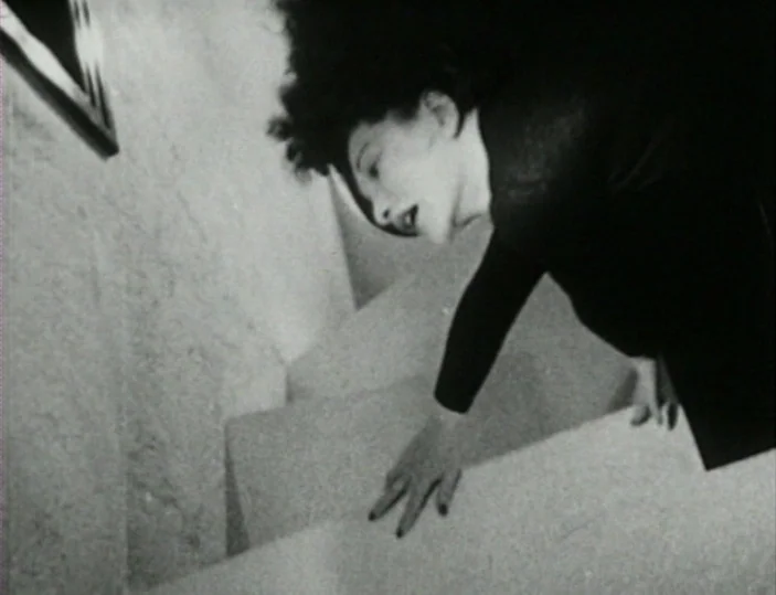 Meshes of the Afternoon (1943) movie still: medium — A dark, angled staircase interior with high-contrast shadows against a pale wall. A woman leans…