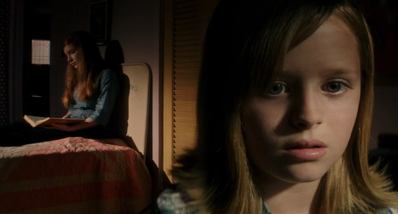 Ouija: Origin of Evil (2016) movie still: medium — A dimly lit bedroom features a girl in the foreground staring directly ahead. Another girl sits…