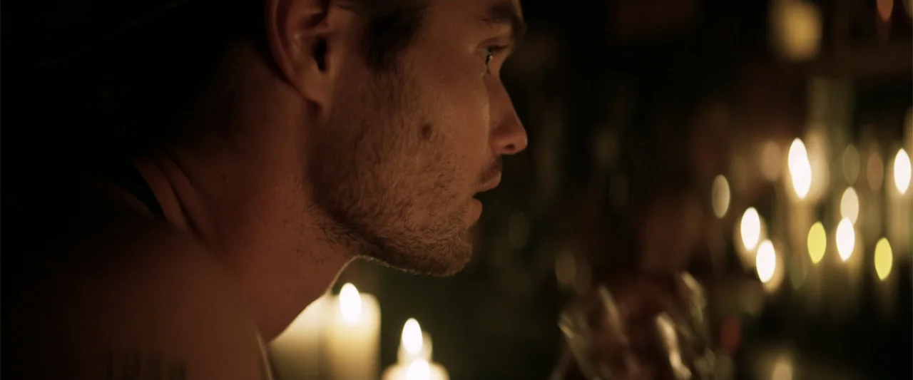 Marked Men: Rule + Shaw (2025) movie still: closeup — A dimly lit space features numerous burning candles arranged on a surface. A man in profile…