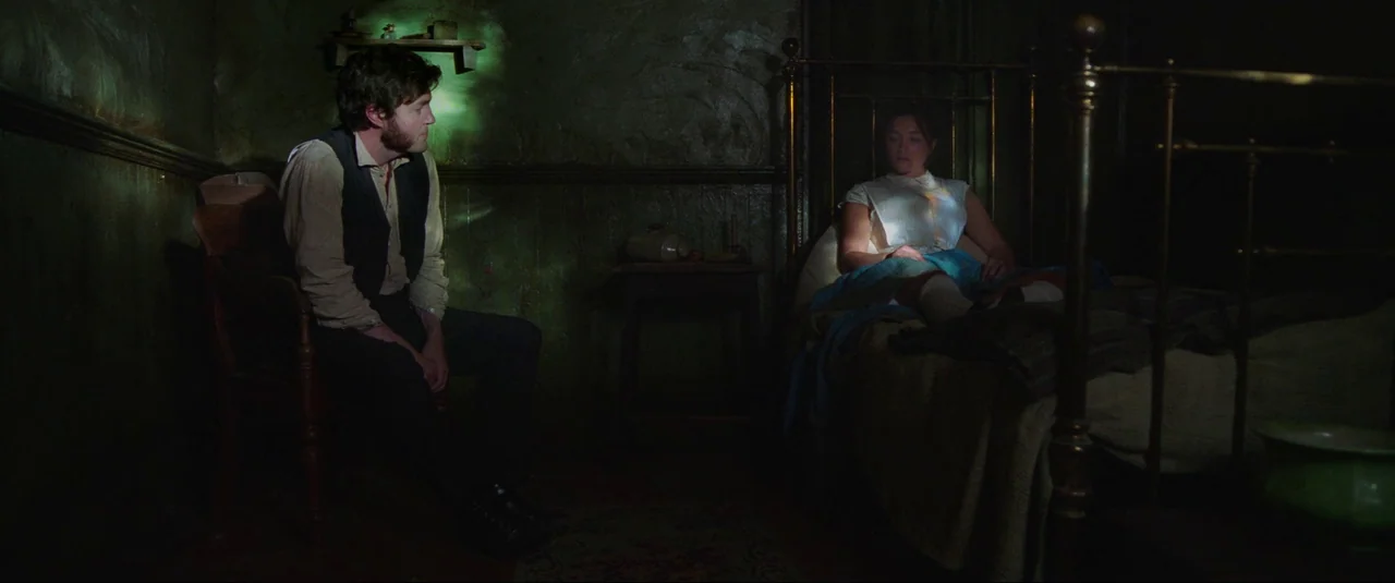 The Wonder (2022) movie still: wide — A dark, small, dimly lit room with rough textured walls and period furniture. A man sits on a wooden chair…