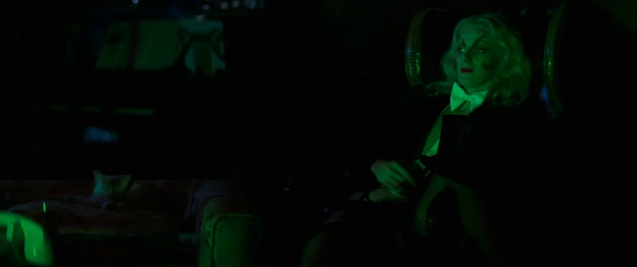 Death Game (1977) movie still: medium — A dark lounge is dominated by a harsh green light source from the side. A woman wearing formal attire sits…