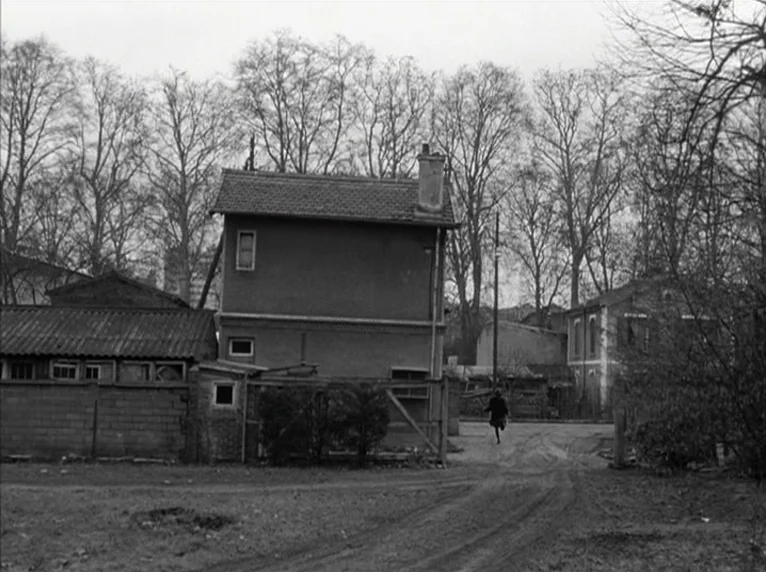Band of Outsiders (Bande à part) (1964) movie still: wide — A quiet residential outskirts area features weathered houses nestled among skeletal,…