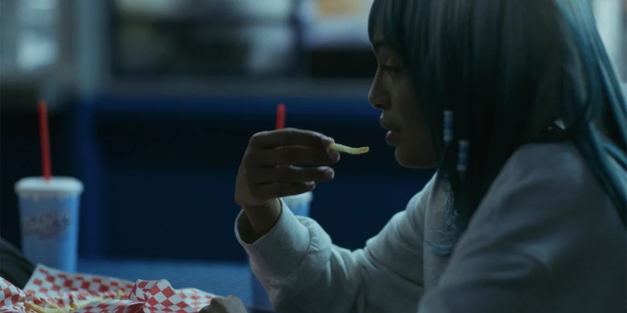 Sitting in Bars with Cake (2023) movie still: medium — A dimly lit fast food establishment with blue-toned surfaces and dark booth seating. A young…
