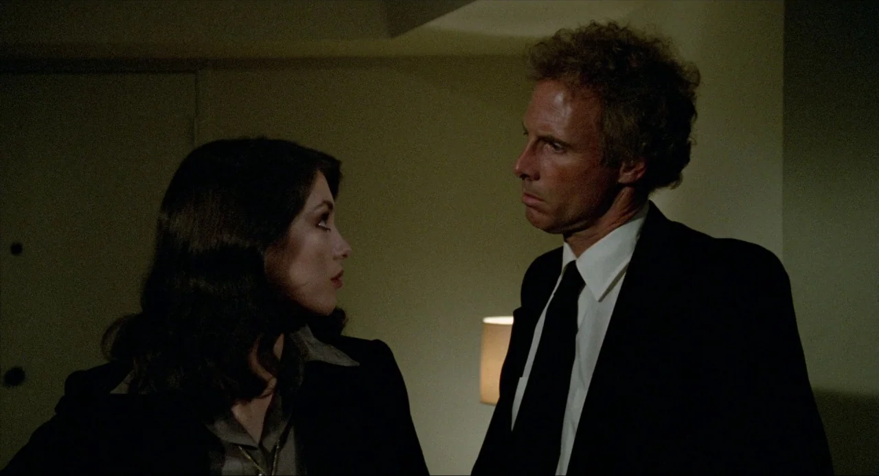 The Driver (1978) movie still: medium — A dimly lit room serves as the setting for two people facing one another. A man in a suit and a woman…