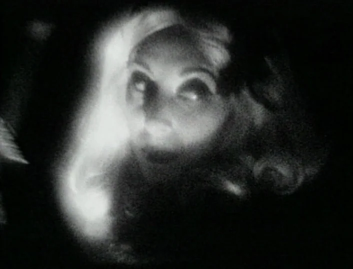 Cowards Bend The Knee (2003) movie still: closeup — A dark, nondescript interior space surrounds a woman appearing from the gloom. Her face is…