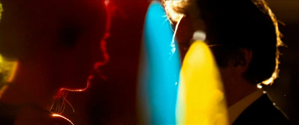 Ocean’s Thirteen (2007) movie still: closeup — A dark, out-of-focus indoor space is illuminated by vibrant neon light sources. Two figures stand in…