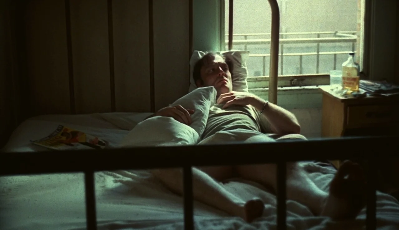 Fat City (1972) movie still: medium — A sparsely furnished room with a metal-frame bed and an open window. A man lies on the mattress, resting his…