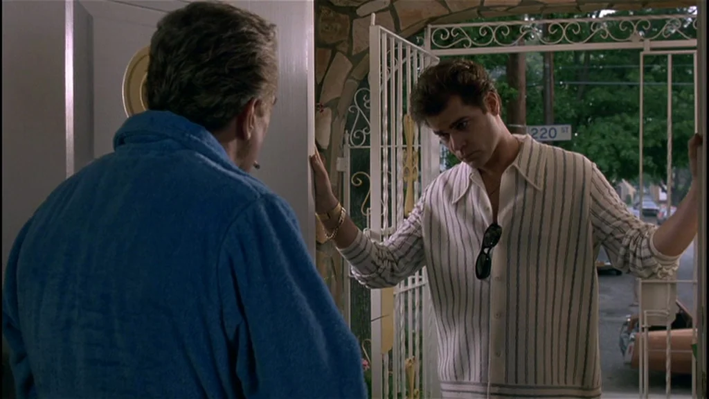 Goodfellas (1990) movie still: medium — A man in a blue robe stands inside a doorway facing an outdoor visitor. The visitor is a man in a striped…