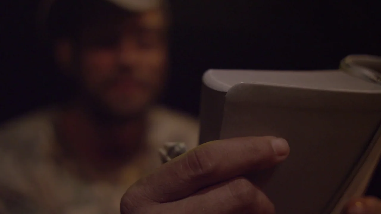 Florida Man (2015) movie still: closeup — A dimly lit interior space with a person partially visible in the background. A foreground hand holds a…