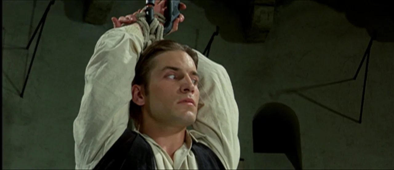 Flesh For Frankenstein (1973) movie still: medium — A stone room with rough masonry walls and dark shadows. A young man is suspended by his hands…