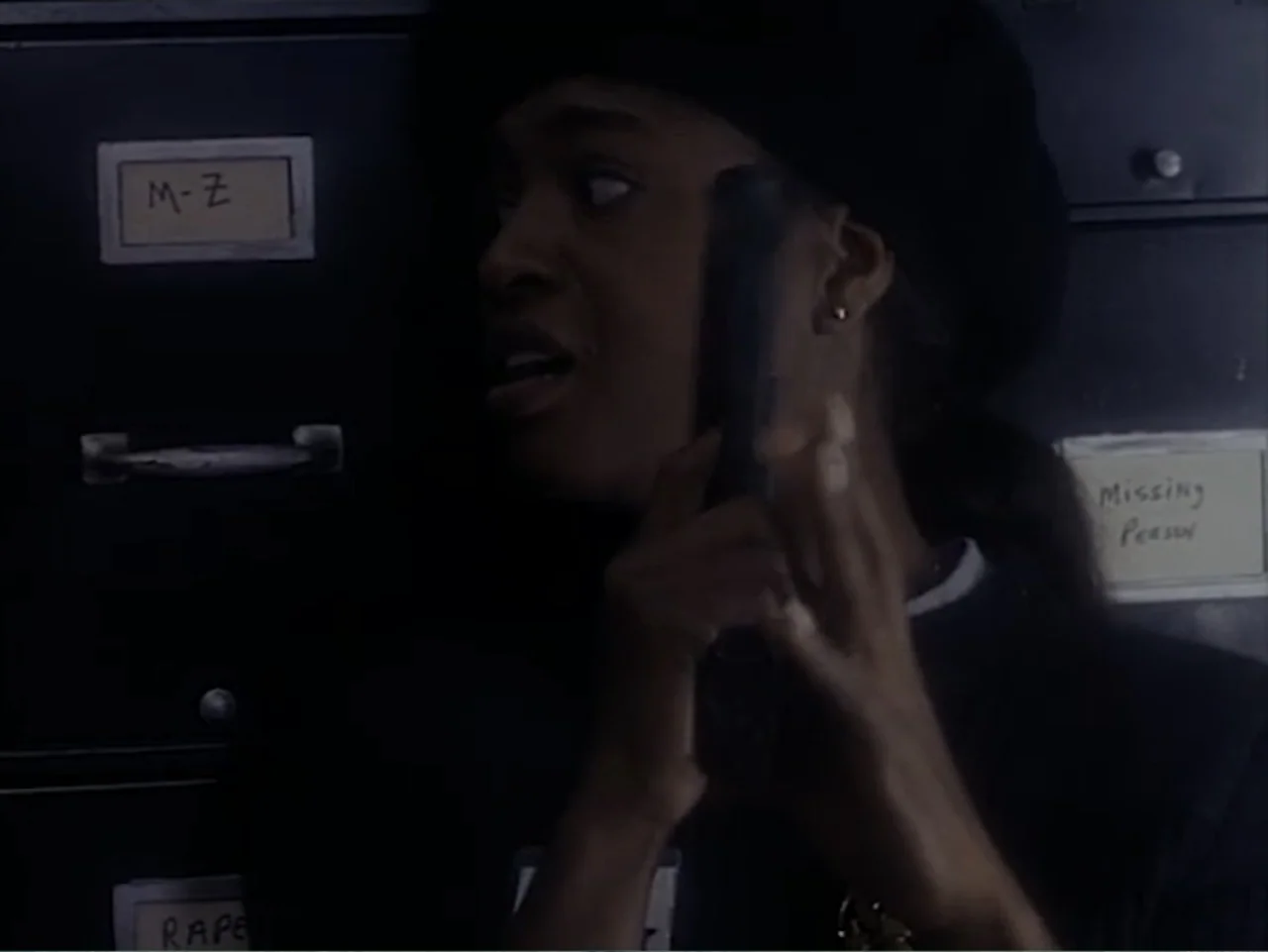 Firepower (1979) movie still: closeup — A dark records room contains rows of black metal filing cabinets with paper labels. A young woman holds a…
