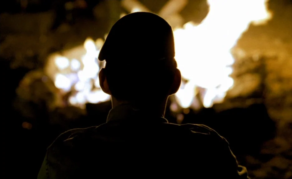 Beau Travail (1999) movie still: medium — A dark outdoor space is illuminated by the intense brightness of a large fire. A person viewed from behind…