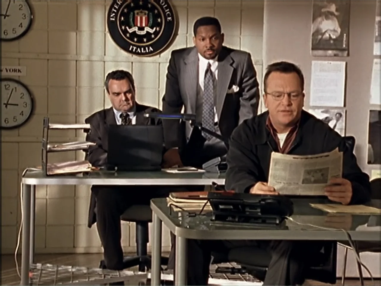 Just 4 Kicks (2003) movie still: medium — An office interior featuring three men working at desks beneath wall clocks and logos. One man reads a…