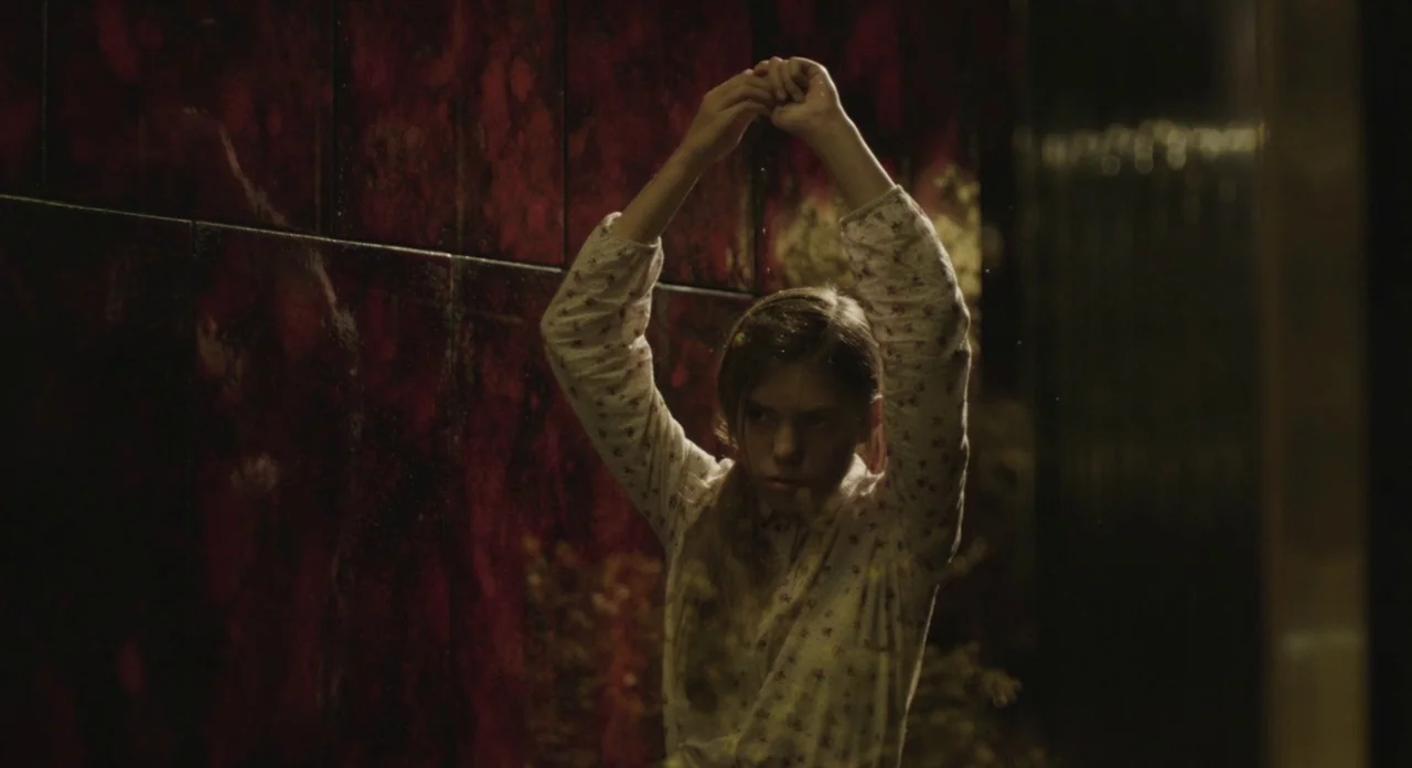 Holy Motors (2012) movie still: medium — A dimly lit room features a wall covered in reflective dark red tiles. A young girl stands with her hands…