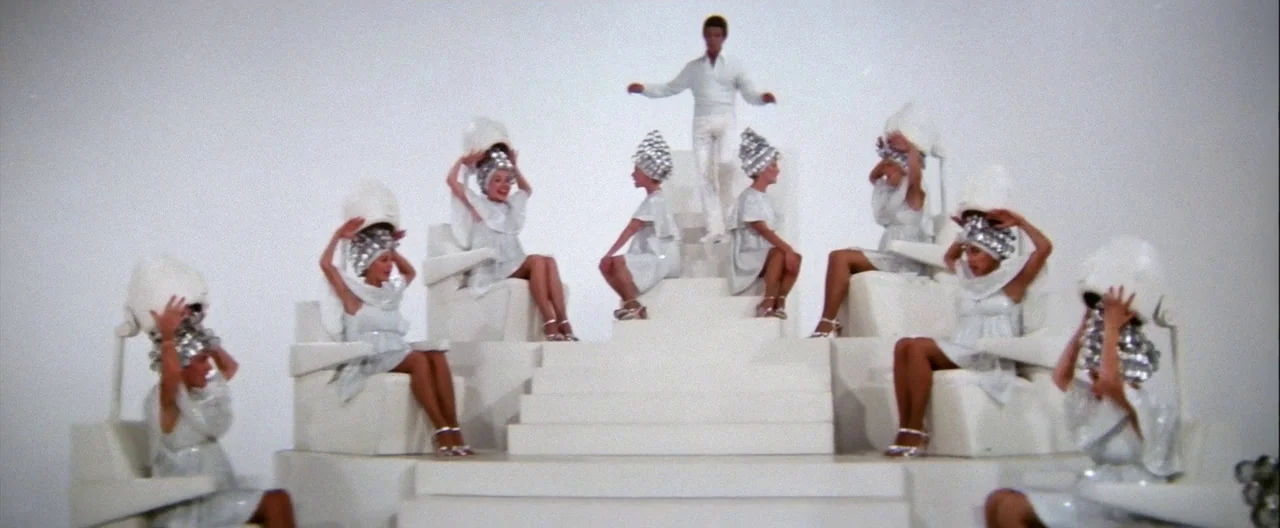 Grease (1978) movie still: wide — A surreal, all-white studio stage features a central tiered platform with white salon chairs. Several women wear…