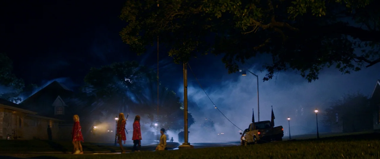 Assassination Nation (2018) movie still: wide — A suburban street at night is blanketed in thick blue fog under street lamps. A small group of…