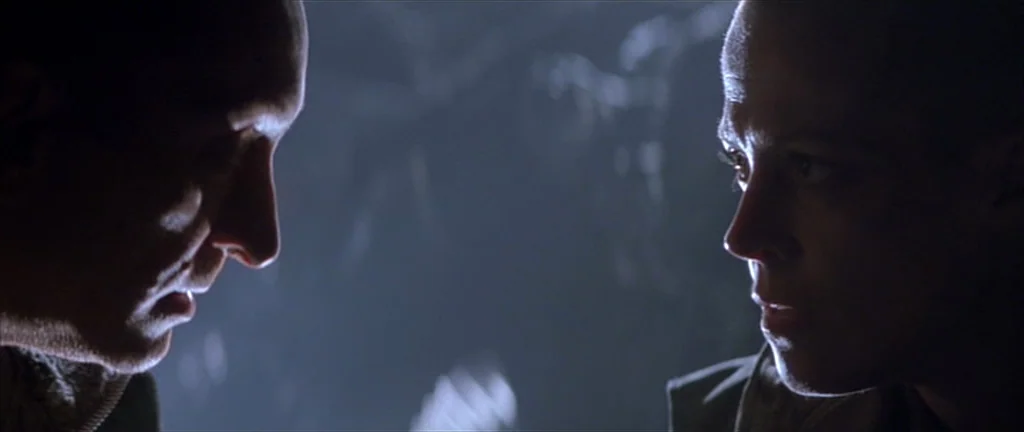 Alien 3 (1992) movie still: closeup — Two individuals stand face-to-face in a dark, indistinct industrial environment. Their faces are illuminated…