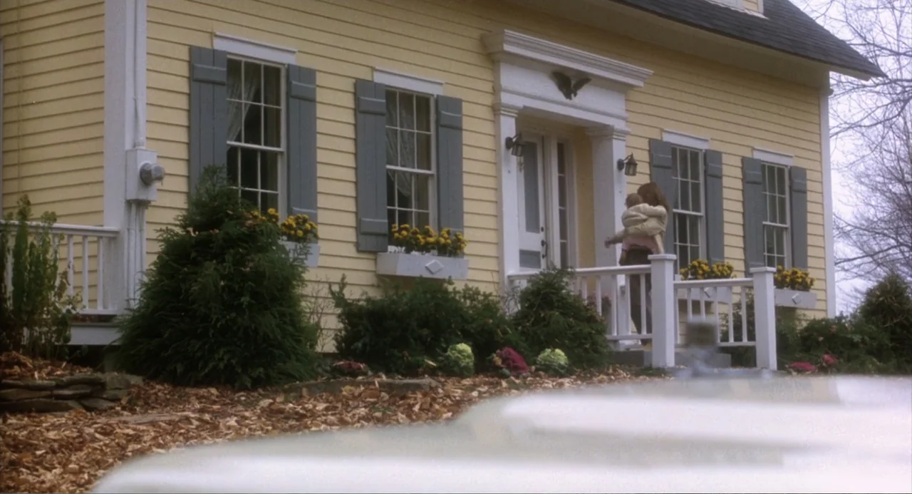 Baby Boom (1987) movie still: wide — A yellow suburban house features white trim, grey shutters, and flower boxes with yellow flowers. A woman…
