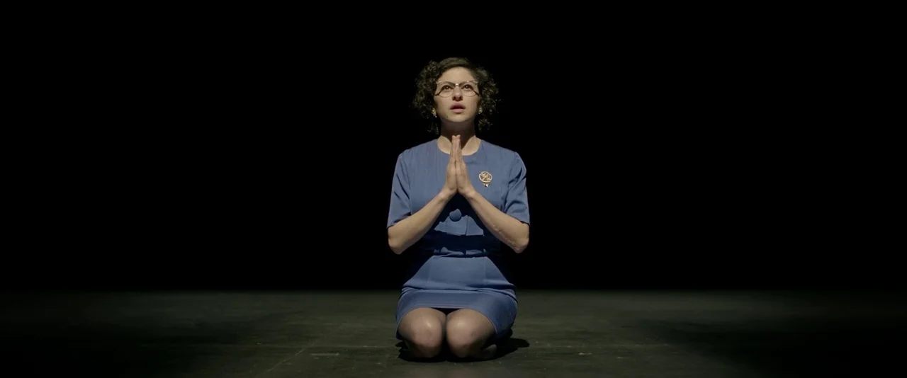 Blaze (2018) movie still: fullbody — A woman kneels in the center of a void with no visible background. She sits on a dark flat surface with her…