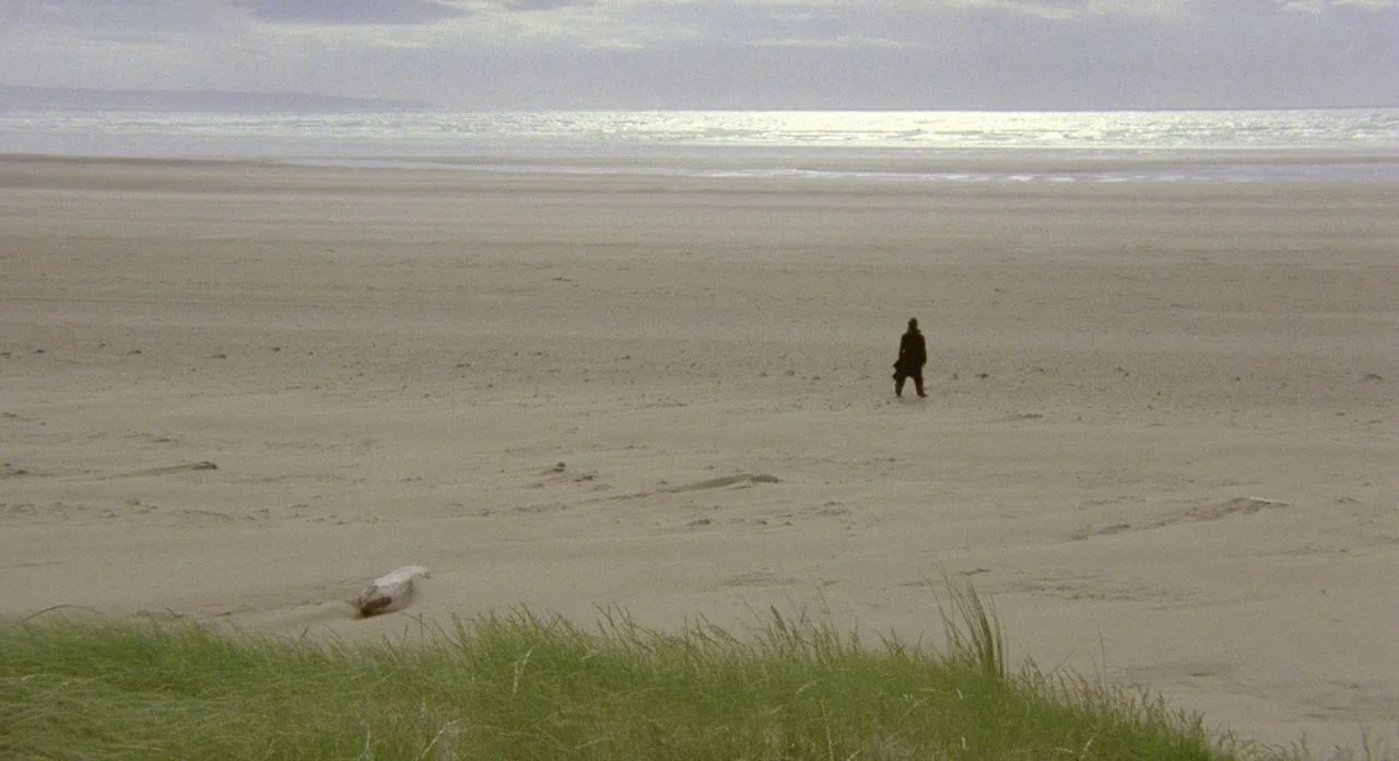 The Shout (1978) movie still: wide — A vast, empty sandy beach stretches toward a distant, reflective ocean under overcast skies. A lone figure in a…