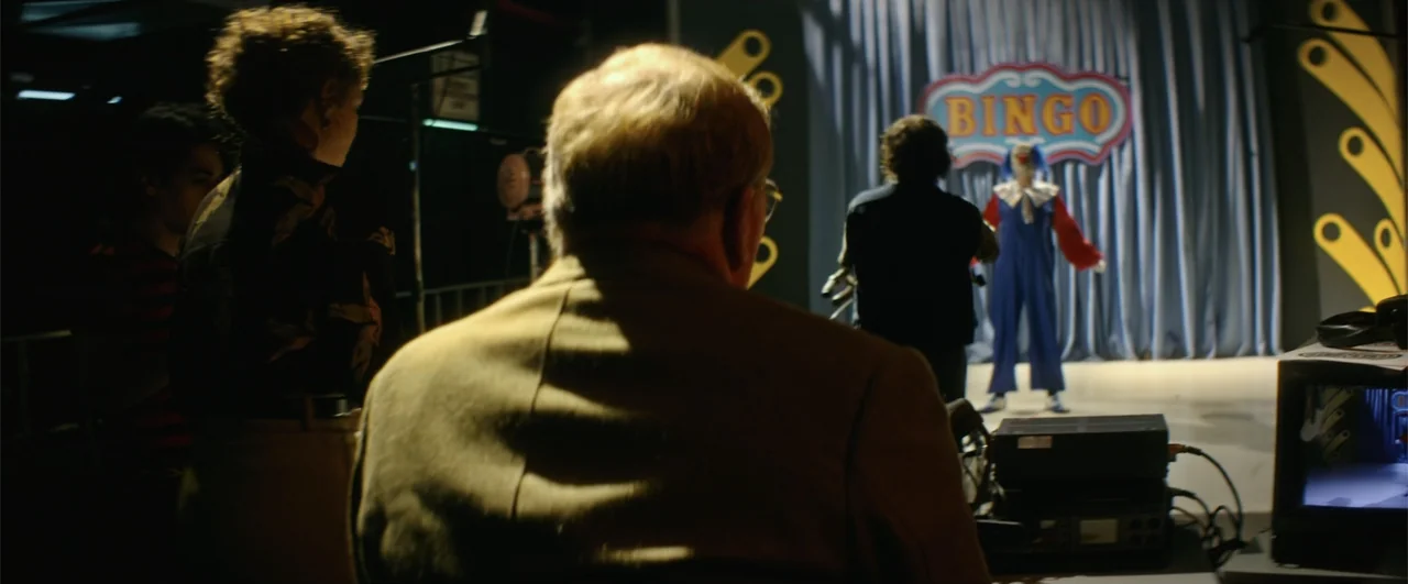 Bingo: The King of the Mornings (2017) movie still: medium — A dark television studio features a brightly lit stage with a bingo sign. A man in a…
