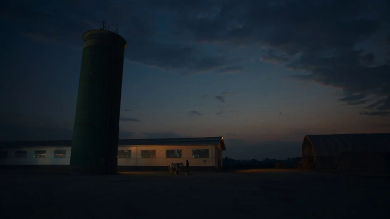 Amanda (2022) movie still: wide — A rural farmstead at dusk features a tall silo and long industrial building. A horse and a human figure are…