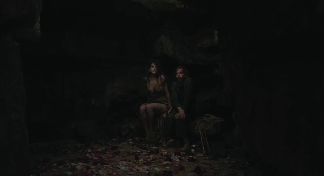 Holy Motors (2012) movie still: Two individuals, a woman in a gold dress and a man in a green robe, sit huddled together within a vast, dark cave,…