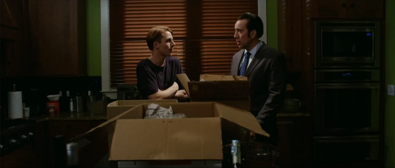 Rage (2014) movie still: medium — A dimly lit kitchen features dark wooden cabinetry and green walls with closed window blinds. Two men stand amidst…