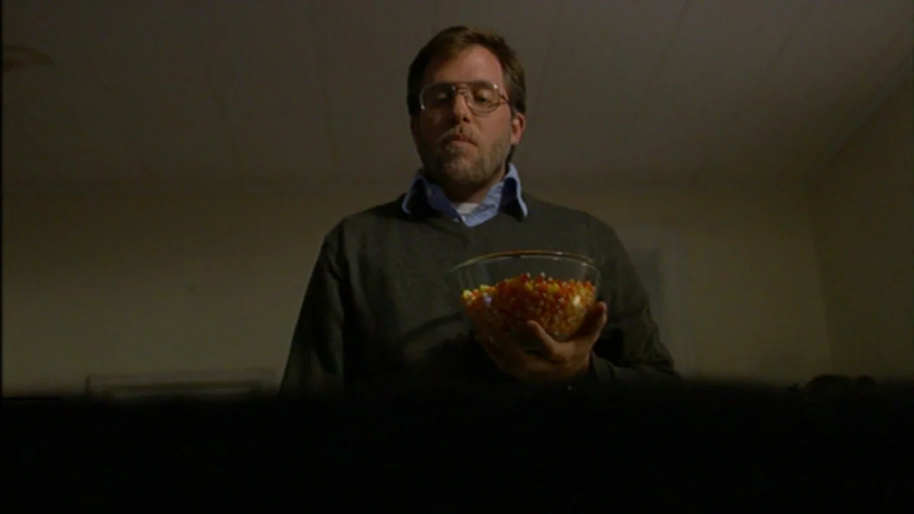 Murder Party movie still: medium — A dimly lit room with an indistinct background wall and ceiling. A man wearing glasses holds a bowl of candy corn.