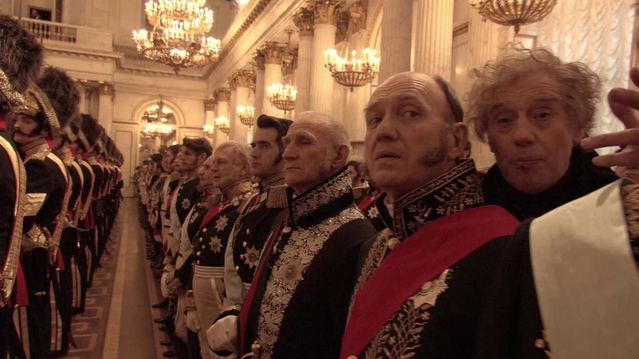 Russian Ark (2002) movie still: medium — A long, opulent hall features white marble columns and multiple glowing gold chandeliers above. A long row…