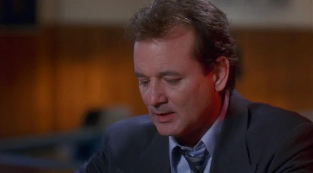 Groundhog Day (1993) movie still: closeup — An interior space with a blurred, warm-toned background indicating an office setting. A man in a suit…